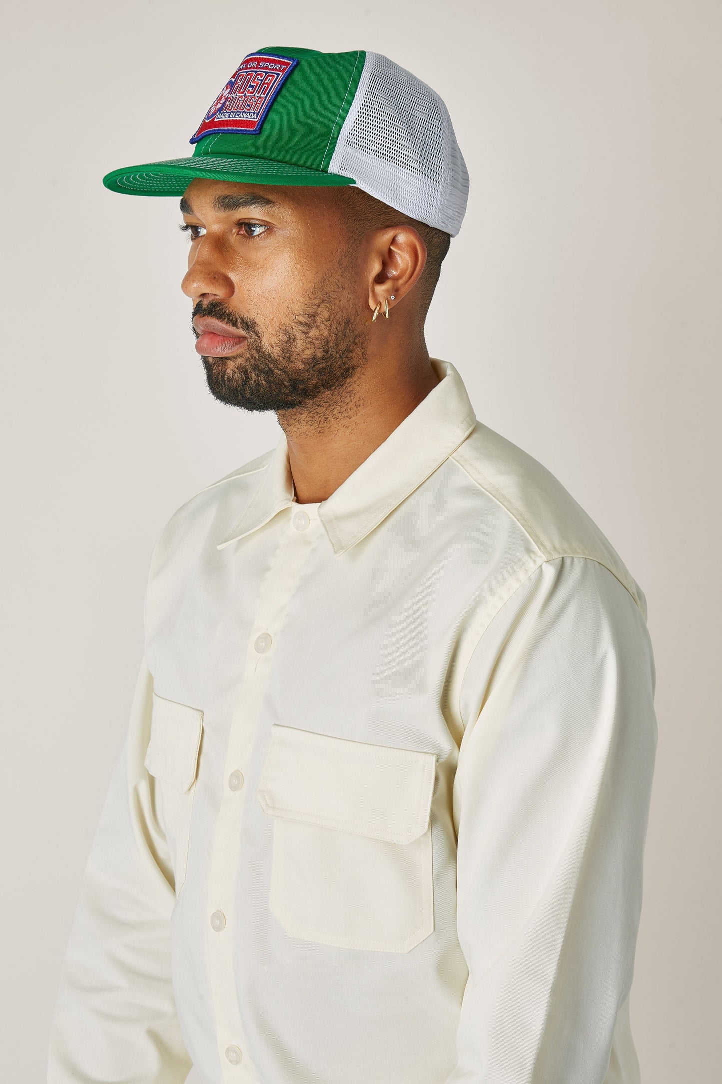 A young bearded man wearing a cream coloured button up long sleeve shirt and a kelly green trucker hat with the red and blue Rosa Rugosa logo patch on the front.