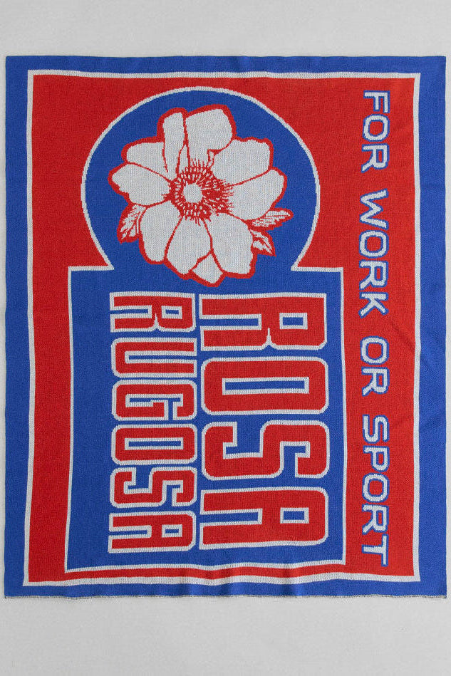 Knitted blanket laid out flat with 'Rosa Rugosa' branding and text on a gray background. The logo is in blue and red. 
